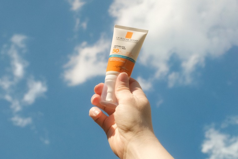 A hand holds tone bottle of sunscreen against a blue sky with clouds