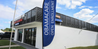 A blue sign reading OBAMACARE OPEN ENROLLMENT is displayed outside a white one-story building.