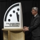 Three people stand to either side of an oversized clock sculpture with text reading "IT IS 85 SECONDS TO MIDNIGHT".