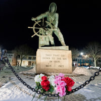 Flowers at the bottom of a memorial statue outside, the plaque on the statue reads "they that go down to the sea in ships"